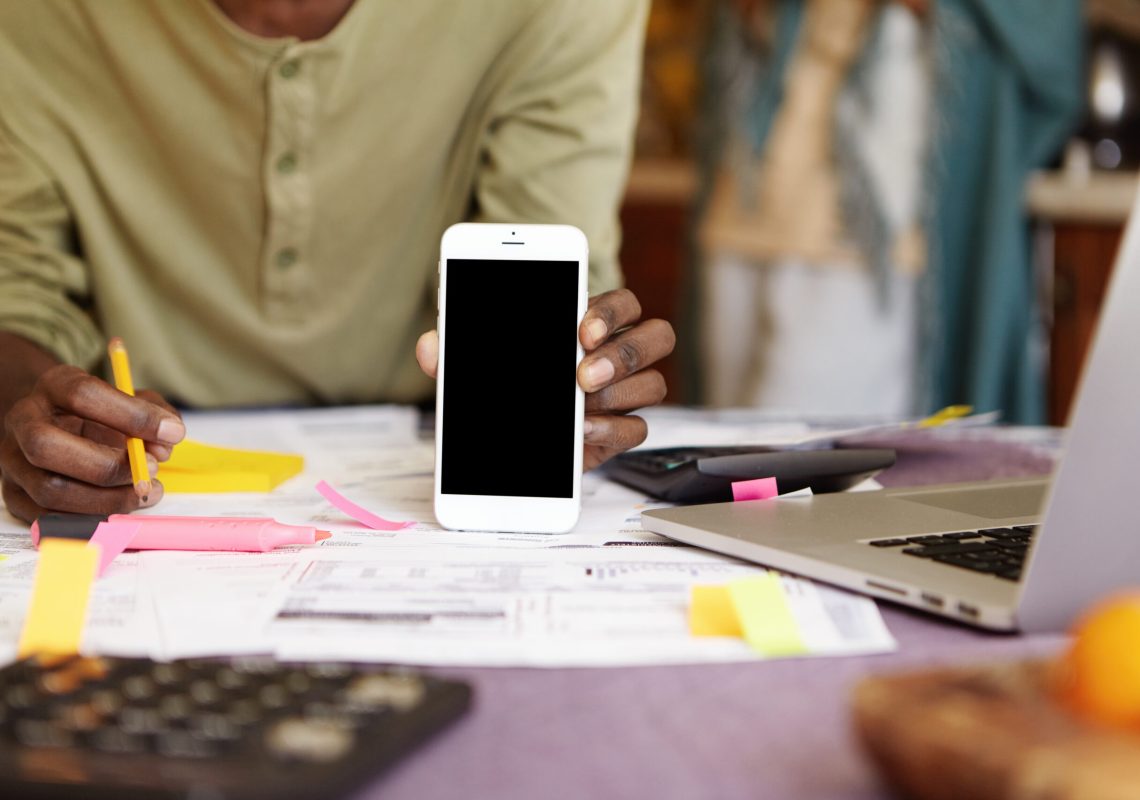 Cropped portrait of dark-skinned man holding black copy space screen cell phone while managing finances and paying bills online, sitting at kitchen table with papers and open laptop computer