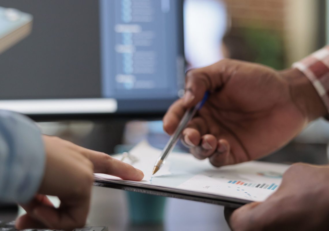 Close up of 3D digital artists reviewing sketch scene plan and simulated render times, Creative industry enthusiasts with CAD open in background discussing technical issues while pointing at clipboard