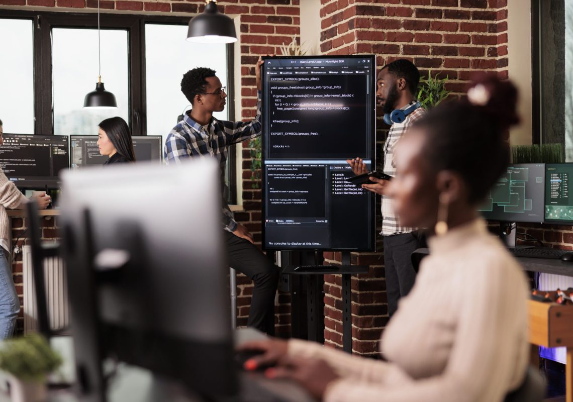 Africann american system engineers analyzing source code looking for errors, checking big tv monitor in it agency office. Software developers working on cyber security innovation in it agency.