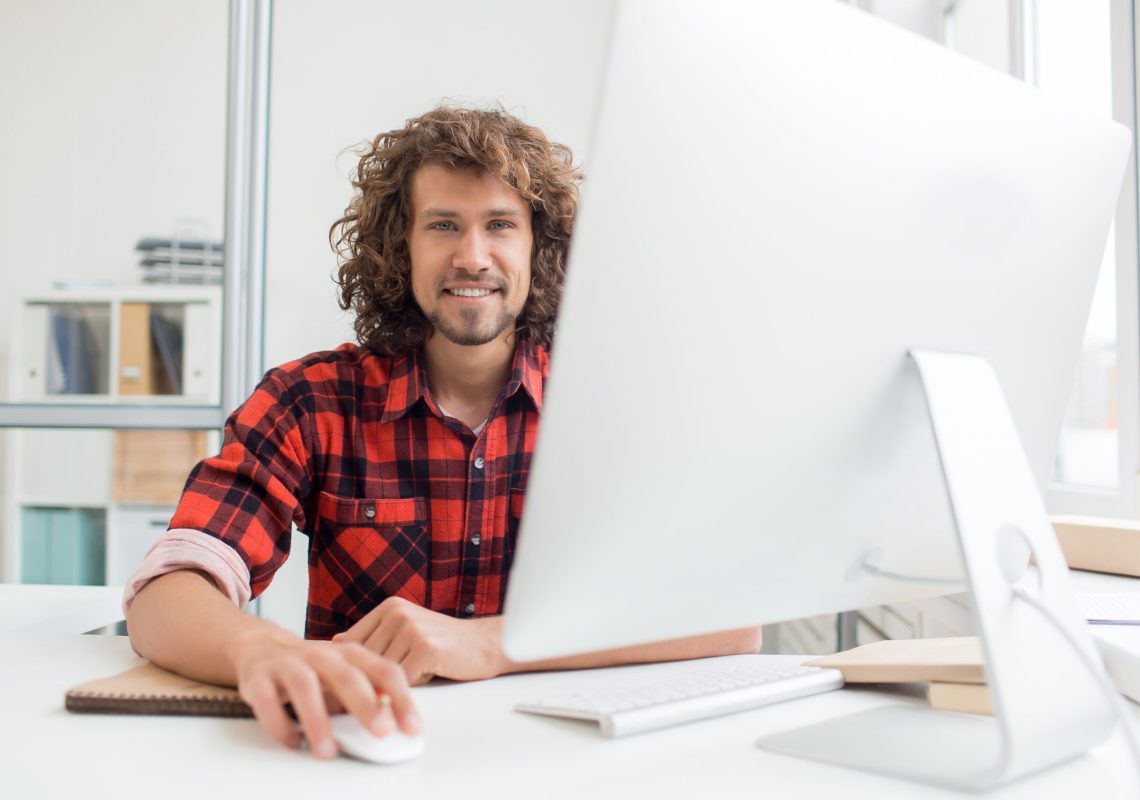 Young designer with curly hair looking at camera with smile while working in front of computer monitor