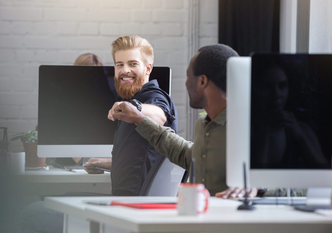 Smiling bearded man giving a fist bump to a male colleague while they are sitting at their computer desks