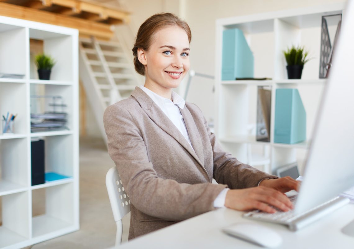 Cheerful young businesswoman computing by workplace while making analysis or research of online data